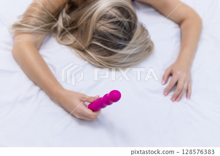 Woman in bed under sheets holding vibrator in hand 128576383