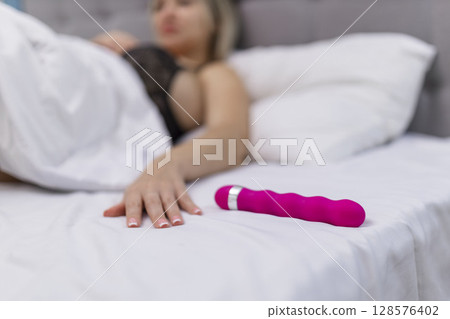 Woman in bed under sheets holding vibrator in hand Woman in bed under sheets holding vibrator in hand 128576402