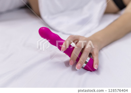 Woman in bed under sheets holding vibrator in hand Woman in bed under sheets holding vibrator in hand 128576404
