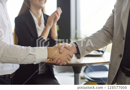 Close up of handshake between business professionals as colleague celebrating success in background. Close up of handshake between business professionals as colleague celebrating success in background. 128577008