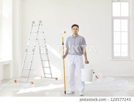 Confident professional painter with paint roller and bucket in hands looking at camera 128577243