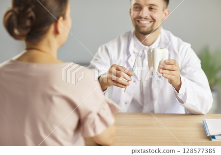 Dentist holding dental toy tooth with toothbrush in hands sitting in dentistry clinic with patient. 128577385