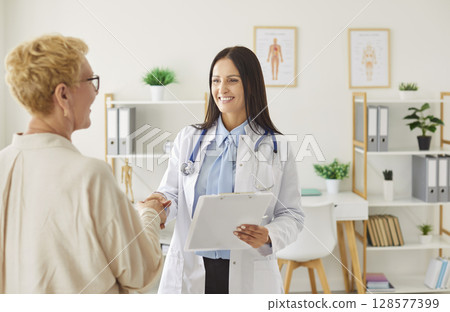 Female doctor smiling and shaking hands with older patient in bright office during consultation. 128577399