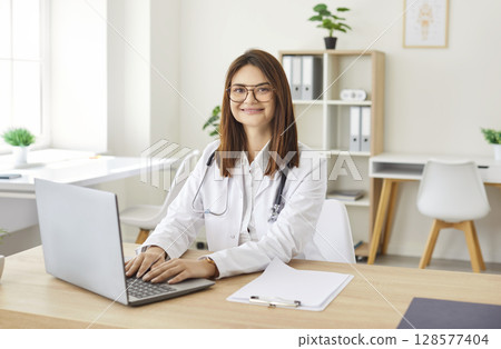 Happy smiling young female GP clinic doctor at laptop, making video call, electronic medical records 128577404