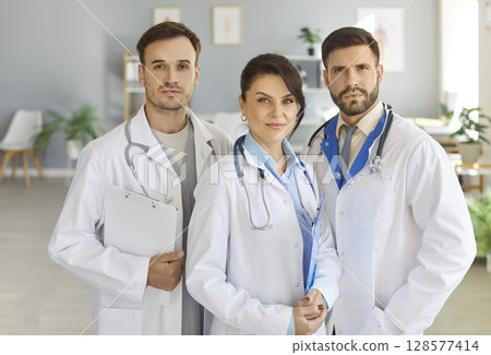 Group of focused professional doctors standing together in medical office and looking at camera. Group of focused professional doctors standing together in medical office and looking at camera. 128577414