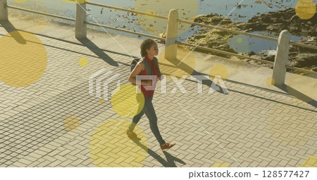 Image of yellow spots over biracial woman walking on promenade 128577427