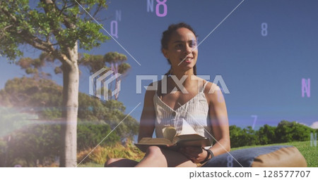 Image of letters and numbers over smiling biracial woman reading in park 128577707