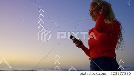 Image of arrows over happy caucasian woman using smartphone at sunset 128577880