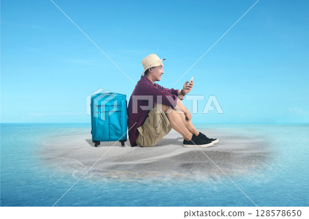 Indonesian southeast asian man using a cellphone sitting with a suitcase on the island with an ocean view and a blue sky. Summer concept 128578650