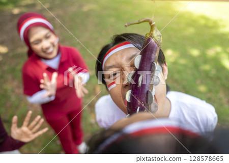 Portrait of an Indonesian southeast asian people celebrate Indonesia Independence Day on the 17th of August with a game of biting coins in eggplant outdoors. Concept of Kemerdekaan Indonesia Portrait of an Indonesian southeast asian people celebrate Indonesia Independence Day on the 17th of August with a game of biting coins in eggplant outdoors. Concept of Kemerdekaan Indonesia 128578665
