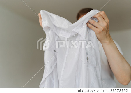 Male wringing out drenched white t-shirt above laundry basket, wet clothing pooling, preparing laundry for washing machine, close-up cropped shot. Concept of household chores and daily routines. Male wringing out drenched white t-shirt above laundry basket, wet clothing pooling, preparing laundry for washing machine, close-up cropped shot. Concept of household chores and daily routines. 128578792