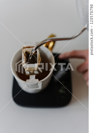 Close-up top view of male hands adjusting kitchen scales during preparing drip coffee, pouring hot water from kettle onto coffee bag on kitchen scales in precise brewing process. 128578798
