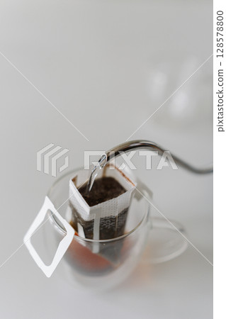 Boiling water pouring from kettle into drip coffee bag placed in glass cup, offering simple and convenient way to brew fresh cup of coffee, close-up. Concept of quick way to enjoy fresh cup of coffee. Boiling water pouring from kettle into drip coffee bag placed in glass cup, offering simple and convenient way to brew fresh cup of coffee, close-up. Concept of quick way to enjoy fresh cup of coffee. 128578800