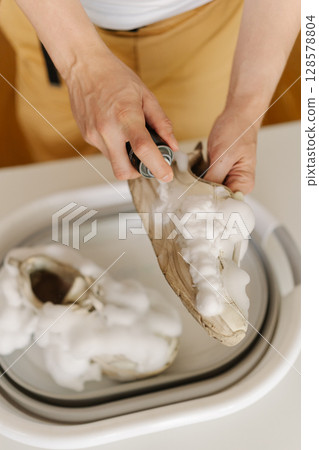 Vertical cropped shot of housewife washing dirty white sneakers with cleaning foam and brush in basin, applying detergent on footwear, doing housework and chores, close-up. 128578804