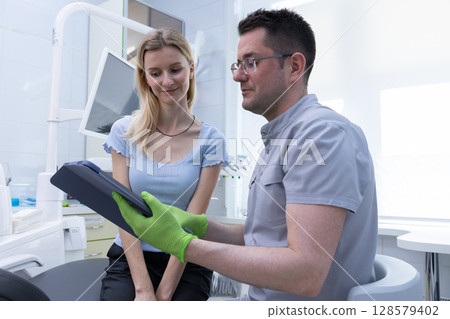In a Modern Dental Clinic, a Dentist With Glasses Discusses Oral Health with a Patient, Showcasing Professionalism and Care in a Brightly Lit Environment 128579402
