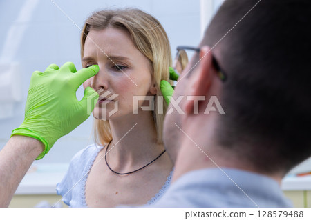 Young Woman Receiving Medical Consultation for Facial Treatment with Professional Practitioner in Health Clinic Setting, Focus on Patient's Expression and Doctor's Hand Preparation 128579488