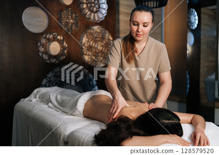 Portrait of female masseuse gently applying massage oil on woman back lying on massage table, providing relaxing and rejuvenating spa treatment in beauty spa salon. Concept of luxury massage. 128579520