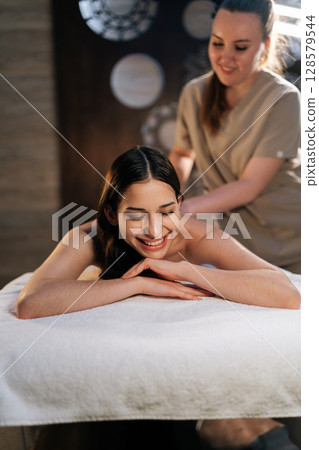 Vertical portrait of cheerful woman client relaxing during back massage in spa salon with dark interior, close-up. Skilled massage therapist providing therapeutic back massage to relaxed female. Vertical portrait of cheerful woman client relaxing during back massage in spa salon with dark interior, close-up. Skilled massage therapist providing therapeutic back massage to relaxed female. 128579544