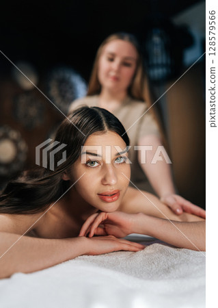 Vertical portrait of attractive female client relaxing with hand massage at beauty spa lying looking at camera. Professional masseur giving back massage to relaxed woman in luxury wellness center. Vertical portrait of attractive female client relaxing with hand massage at beauty spa lying looking at camera. Professional masseur giving back massage to relaxed woman in luxury wellness center. 128579566
