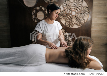 Wide shot of female massage therapist working on client woman back, delivering soothing treatment in spa salon with softly illuminated wellness environment. Concept of rest with spa treatment Wide shot of female massage therapist working on client woman back, delivering soothing treatment in spa salon with softly illuminated wellness environment. Concept of rest with spa treatment 128579568