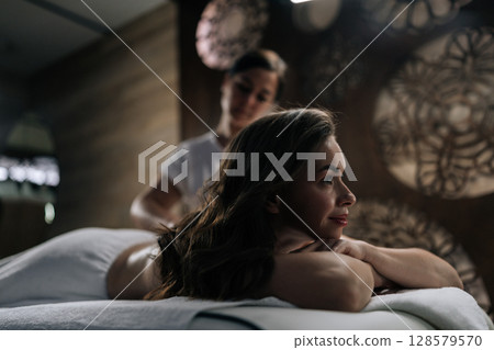 Side view of serene young woman relaxing during back massage in spa with soft light. Beautiful naked female with perfect skin getting relaxing massage. Concept of healthy lifestyle and body care Side view of serene young woman relaxing during back massage in spa with soft light. Beautiful naked female with perfect skin getting relaxing massage. Concept of healthy lifestyle and body care 128579570