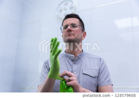 Professional Healthcare Worker Putting on Green Medical Gloves in a Clean Clinic Environment, Ready for Treatment, With a Clock in the Background Indicating Time During Procedures 128579608