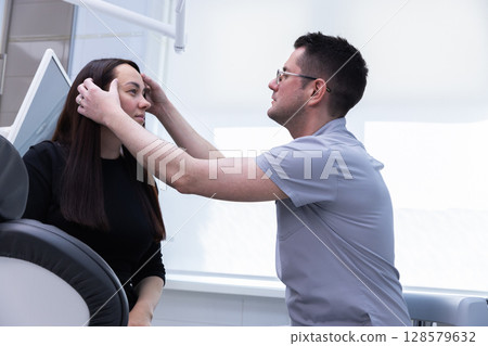 A dental professional examining a patient in a modern clinic setting, focusing on facial features and dental health with a gentle and caring approach during the consultation process. 128579632