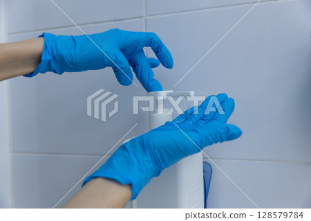 Person with blue latex gloves dispensing liquid soap or sanitizer from a pump bottle, emphasizing cleanliness and hygiene practices in a modern bathroom or kitchen setting. 128579784