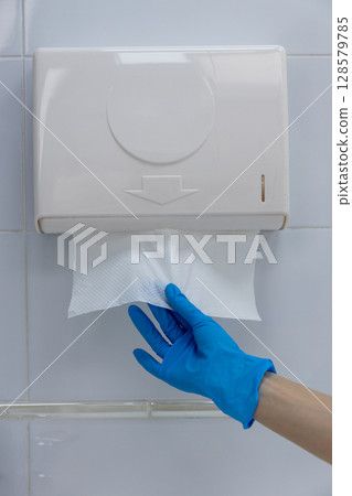 Hand Reaching for Paper Towel from Wall-Mounted Dispenser with Blue Gloves in Clean Restroom Hand Reaching for Paper Towel from Wall-Mounted Dispenser with Blue Gloves in Clean Restroom 128579785