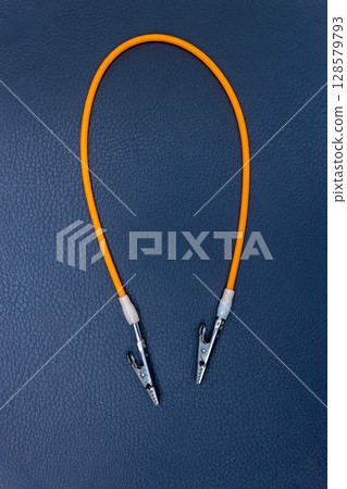 A Close-up View of an Orange Electrical Wire with Alligator Clips on Either End, Set Against a Textured Dark Blue Background A Close-up View of an Orange Electrical Wire with Alligator Clips on Either End, Set Against a Textured Dark Blue Background 128579793