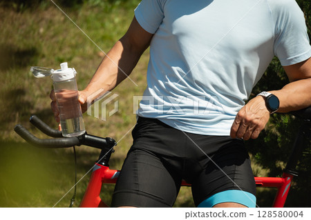 Man resting on bicycle holding water bottle at urban public park. Relaxation and refreshment outdoors among greenery. Take a break. Outdoor summer activity. Man resting on bicycle holding water bottle at urban public park. Relaxation and refreshment outdoors among greenery. Take a break. Outdoor summer activity. 128580004