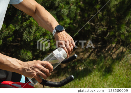 Close up of hands of cyclist check apps performance data by smartwatch and holding water bottle, standing with his bike in park on sunny day in forest. Close up. 128580009
