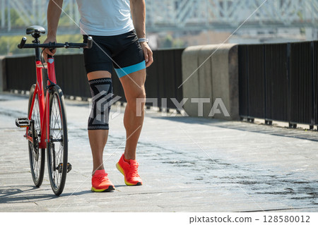 Cyclist walks with bandage on knee at public park. Unhappy cyclist with knee pain and injury outdoors. Cyclist walks with bandage on knee at public park. Unhappy cyclist with knee pain and injury outdoors. 128580012