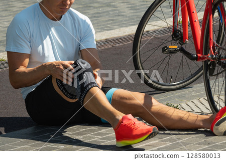 Cyclist fell off his bike and injured his knee. Unhappy cyclist with knee pain and injury outdoors. Close up. 128580013