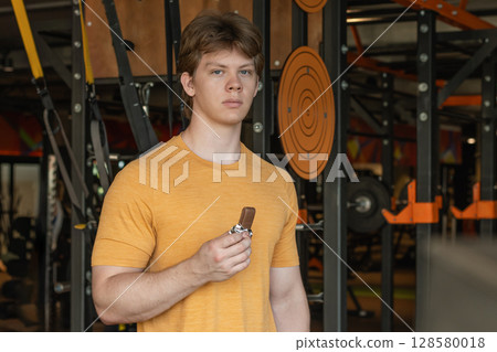 Sporty man eating energy bar. Muscular athlete snacks protein bar while resting after workout at gym or fitness center. Sporty man eating energy bar. Muscular athlete snacks protein bar while resting after workout at gym or fitness center. 128580018