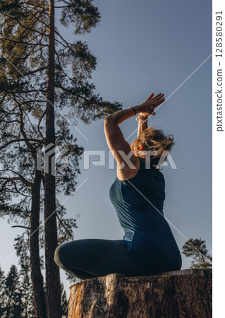 A woman practicing yoga performs a sitting in lotus position on a tree stump, meditating alone in casual clothes on a sunny autumn day in the park A woman practicing yoga performs a sitting in lotus position on a tree stump, meditating alone in casual clothes on a sunny autumn day in the park 128580291
