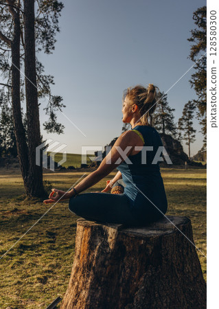 A woman practicing yoga performs a sitting in lotus position on a tree stump, meditating alone in casual clothes on a sunny autumn day in the park A woman practicing yoga performs a sitting in lotus position on a tree stump, meditating alone in casual clothes on a sunny autumn day in the park 128580300