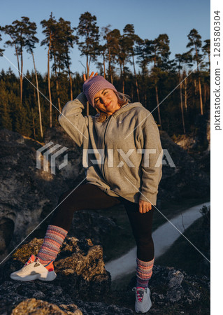 A woman in sportswear sitting on a rock at sunset, wearing a hat and looking like a hiker. The warm summer light and natural setting evoke themes of adventure, travel, and outdoor freedom. 128580304