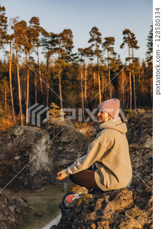 A woman in sportswear sitting on a rock at sunset, wearing a hat and looking like a hiker. The warm summer light and natural setting evoke themes of adventure, travel, and outdoor freedom. 128580334