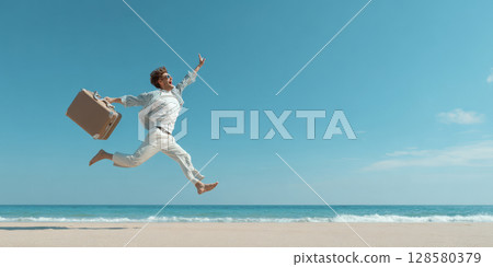 A dynamic image of a man mid-air, leaping with a suitcase on a white sand beach against a clear blue sky, calm ocean, representing the excitement and happiness of reaching a dream vacation destination 128580379