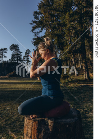 A woman practicing yoga performs a sitting in lotus position on a tree stump, meditating alone in casual clothes on a sunny autumn day in the park A woman practicing yoga performs a sitting in lotus position on a tree stump, meditating alone in casual clothes on a sunny autumn day in the park 128580586
