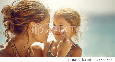 A loving mother applies sunscreen to her child's face at the beach, ensuring sun safety. Perfect for themes of summer, family, skincare, and protection 128580672