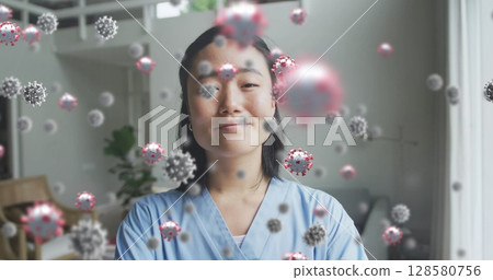 Young Asian woman wearing medical scrubs in living room featuring floating 3D coronavirus models 128580756