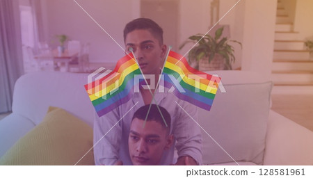 Image of rainbow flags over happy biracial gay couple sitting on sofa at home 128581961