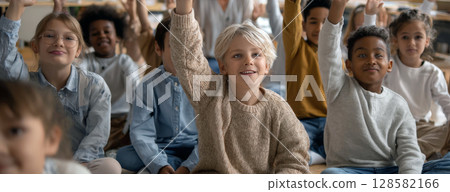 A vibrant group of diverse students, seated attentively in a classroom, eagerly raise their hands, showcasing their enthusiasm and active participation in learning 128582166