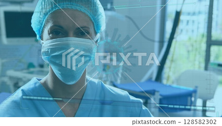 Standing worker wearing blue scrubs, cap and mask in treatment room, with monitors, virus overlay 128582302