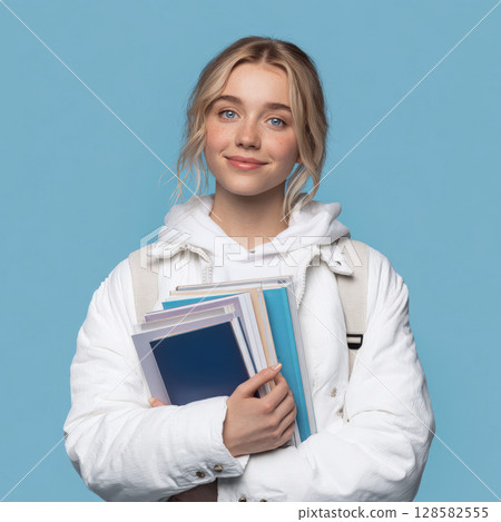 Portrait of a smiling young woman holding books, ideal for themes of education, studying, college life, and back-to-school marketing. Posed against a clean blue background 128582555