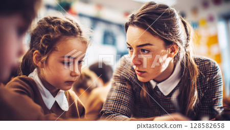 A dedicated female teacher, looking intently, leans in to assist a young female student who is focused on her work, illustrating a moment of personalized instruction in a classroom setting 128582658