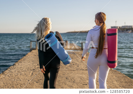 Yoga Women Beach Pier - Two women walk on a pier with yoga mats, heading towards the sea for a workout. Yoga Women Beach Pier - Two women walk on a pier with yoga mats, heading towards the sea for a workout. 128585720