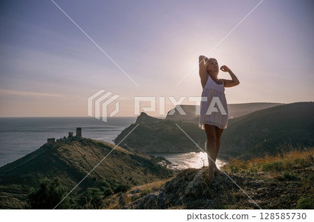A blonde woman stands on a hill overlooking the ocean. She is wearing a white dress and she is enjoying the view. A blonde woman stands on a hill overlooking the ocean. She is wearing a white dress and she is enjoying the view. 128585730